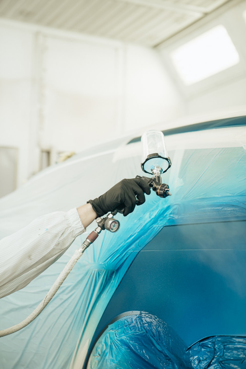 Painter in protective gear sprays blue car with spray gun.