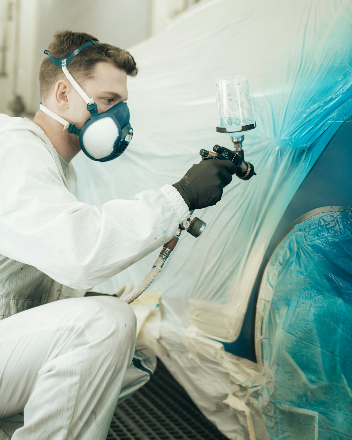 Man in protective gear sprays blue paint on car part.