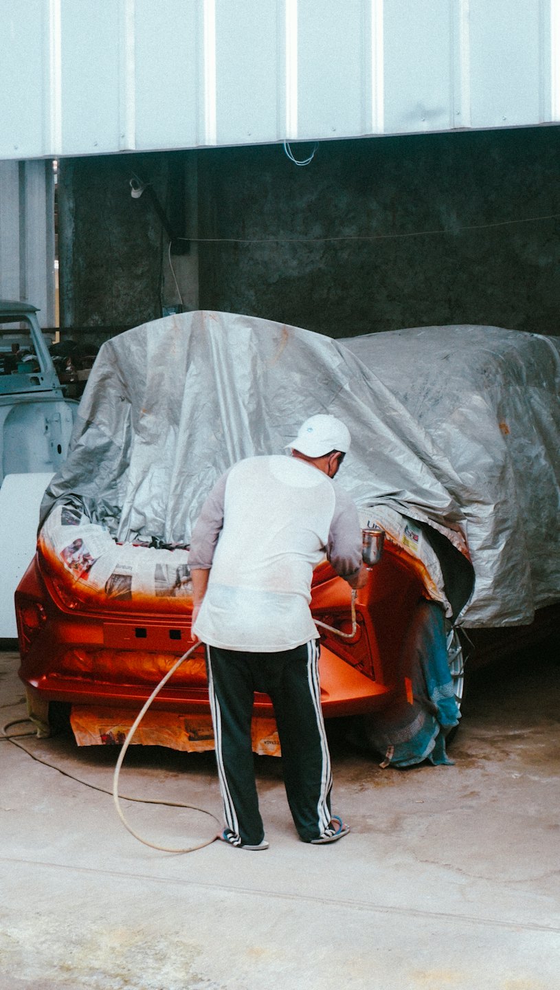 An auto painter sprays the bumper of a car inside a workshop.